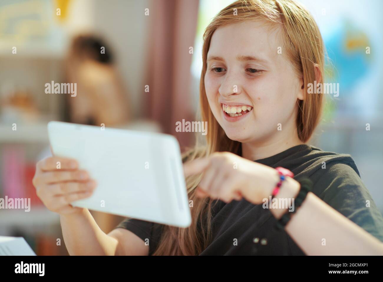 Jeune fille d'école moderne souriante en chemise grise avec tablette PC apprentissage à distance à la maison par temps ensoleillé. Banque D'Images