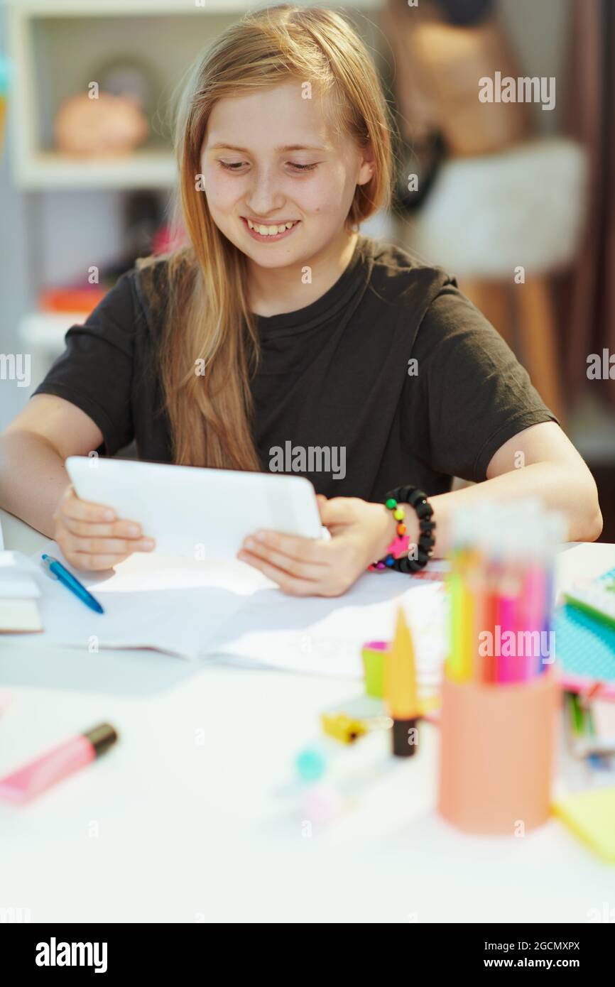 Jeune fille d'école moderne souriante en chemise grise avec tablette PC ayant l'éducation à distance à la maison par beau temps. Banque D'Images