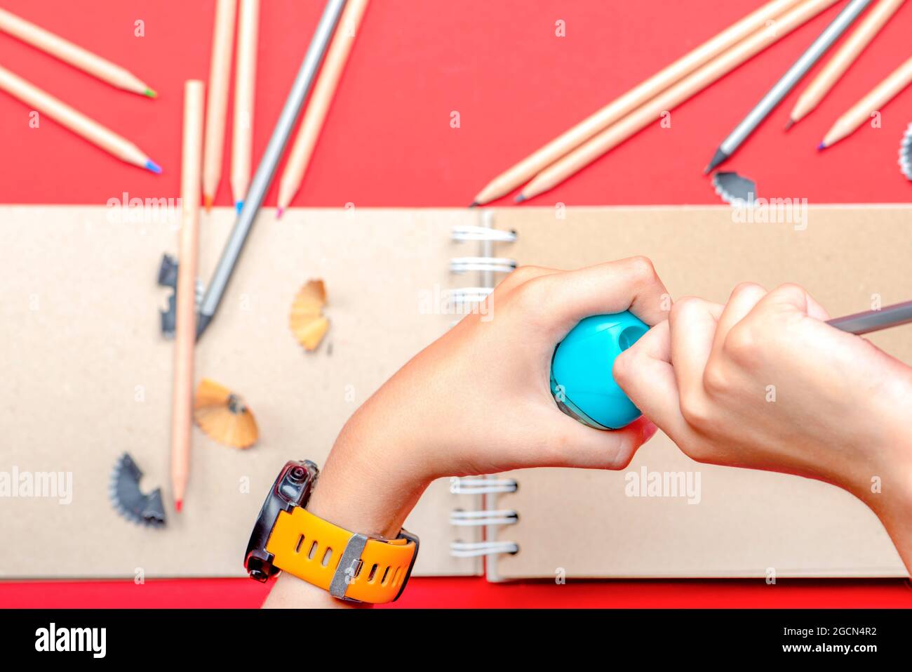 Aiguisez les crayons. Les mains de l'enfant aiguisent un crayon à l'école dans la leçon. En arrière-plan, un carnet de croquis et des crayons de couleur sur la table. Rouge Banque D'Images