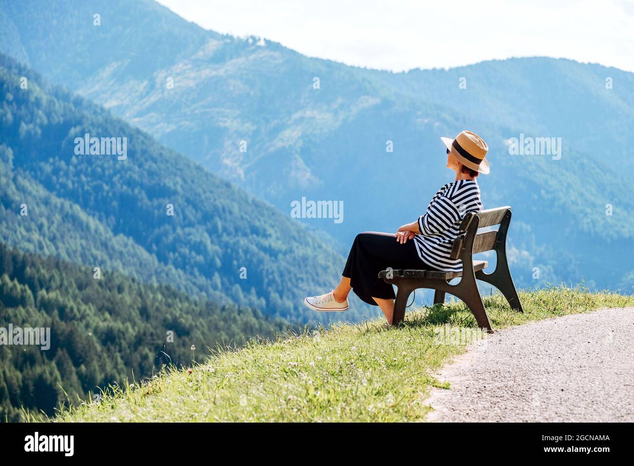 Jeune femme habillée à la mode avec chapeau de paille, assise sur un banc, avec vue sur le village de Santa Maddalena et superbes pics pittoresques des Alpes Dolomites Banque D'Images