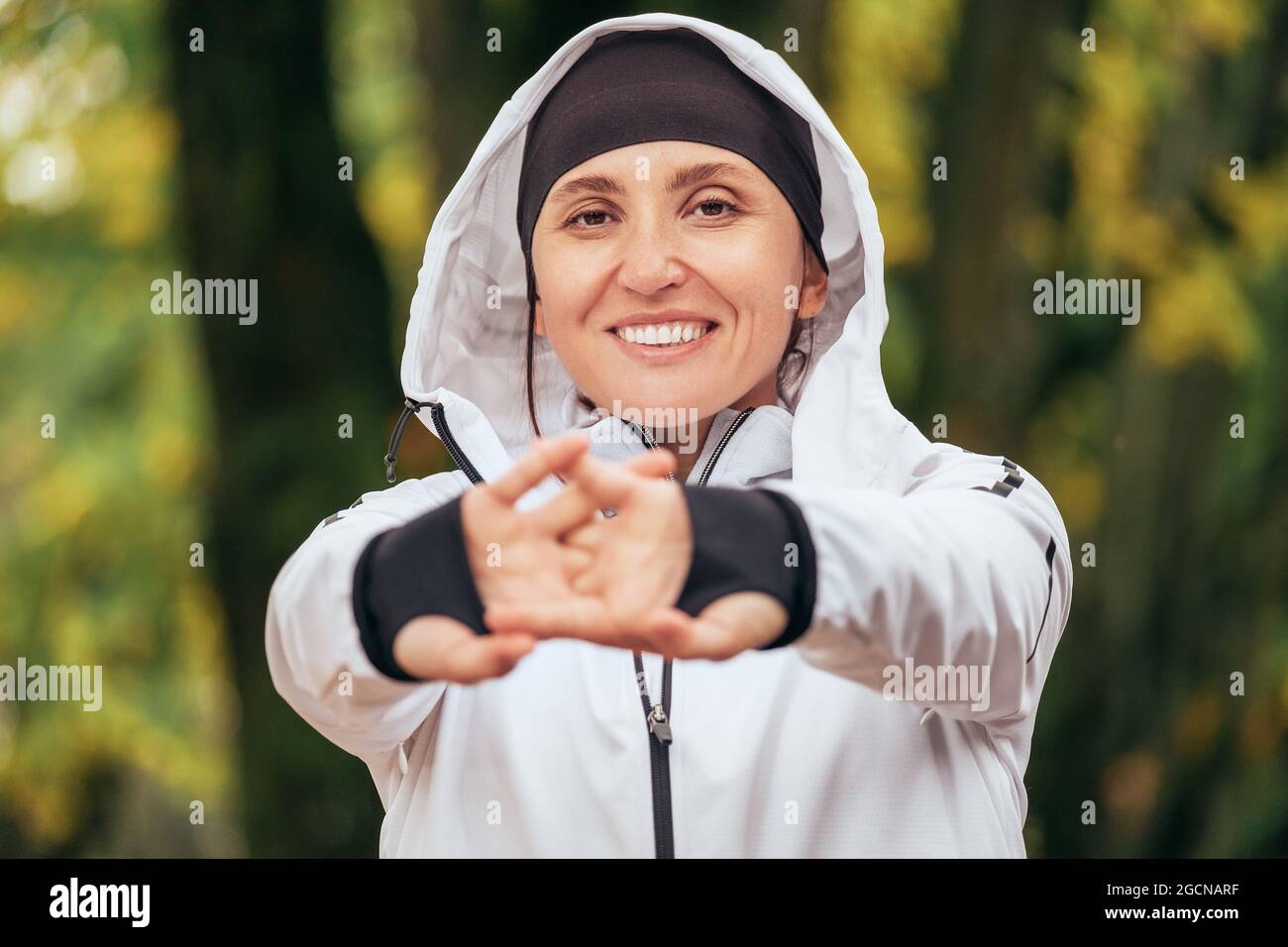 Une femme sportive Smiling Fit portant une veste de course à pied moderne et imperméable qui s'étire les poignets et les doigts dans un parc d'automne vert jaune coloré. spo actif Banque D'Images