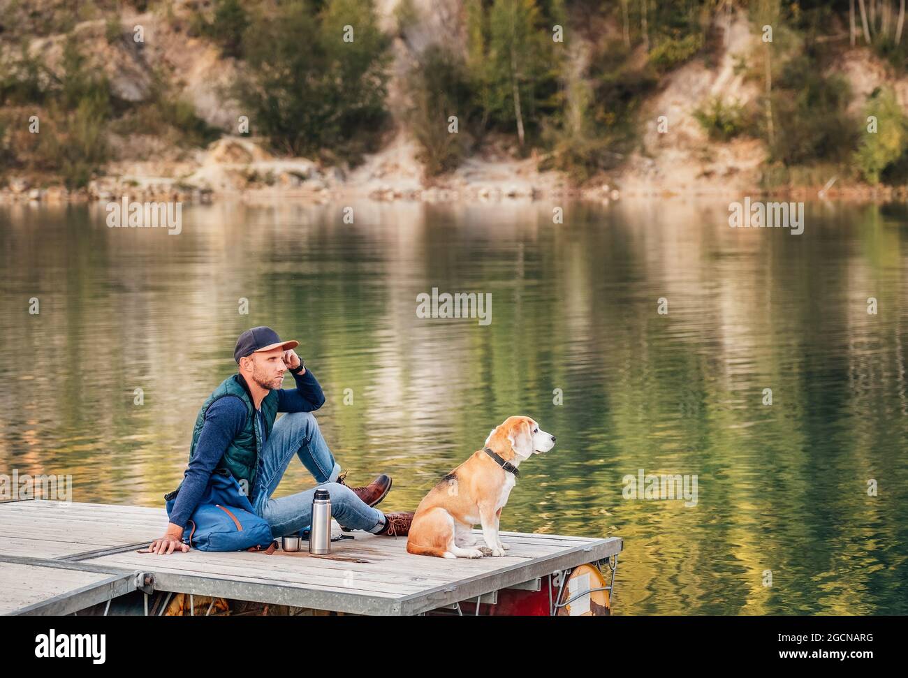 Homme maître de chien et son ami beagle assis sur la jetée en bois sur le lac de montagne et appréciant le paysage pendant leur marche dans les mers d'automne Banque D'Images