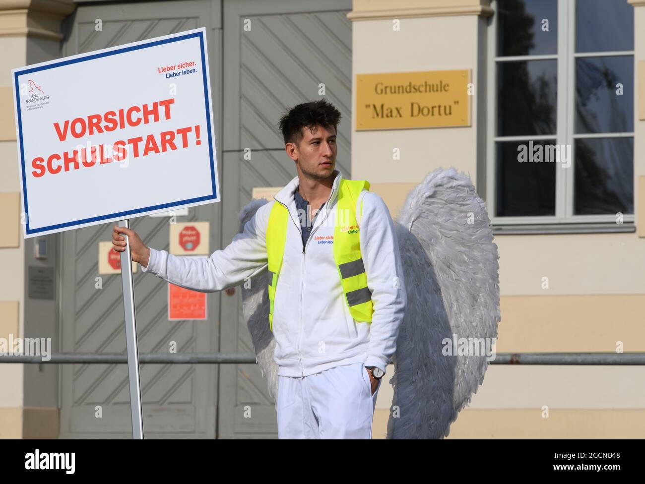 Potsdam, Allemagne. 09e août 2021. Un jeune homme costumé comme un ange de la campagne de sécurité routière se trouve sur la route devant l'école primaire Max-Dortu avec le panneau 'Beware le début de l'école'. Dans les quarante premiers jours après le début de l'école après les vacances d'été, la police veut augmenter les contrôles de la circulation aux heures de priorité. Dans le même temps, la campagne de sécurité routière "Lieber sicher. Lieber leben.' a été lancé pour promouvoir la prise en considération parmi les cyclistes et les automobilistes et pour éduquer les enfants et les parents sur la sécurité routière. Credit: Soeren Stache/dpa-Zentralbild/dpa/Alay Live News Banque D'Images