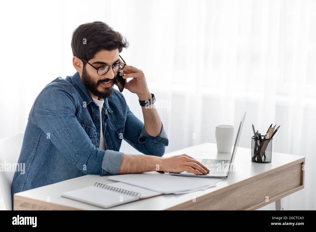 Travailler à domicile. Indépendant arabe, homme parlant sur téléphone portable tout en travaillant avec des papiers et un ordinateur portable, assis au bureau à domicile Banque D'Images