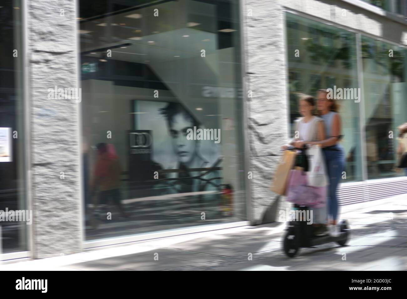 E-mobilité à Munich.Deux femmes sur e scooter avec bagages, sacs de papier suspendus sur guidon.Mouvement flou. Banque D'Images