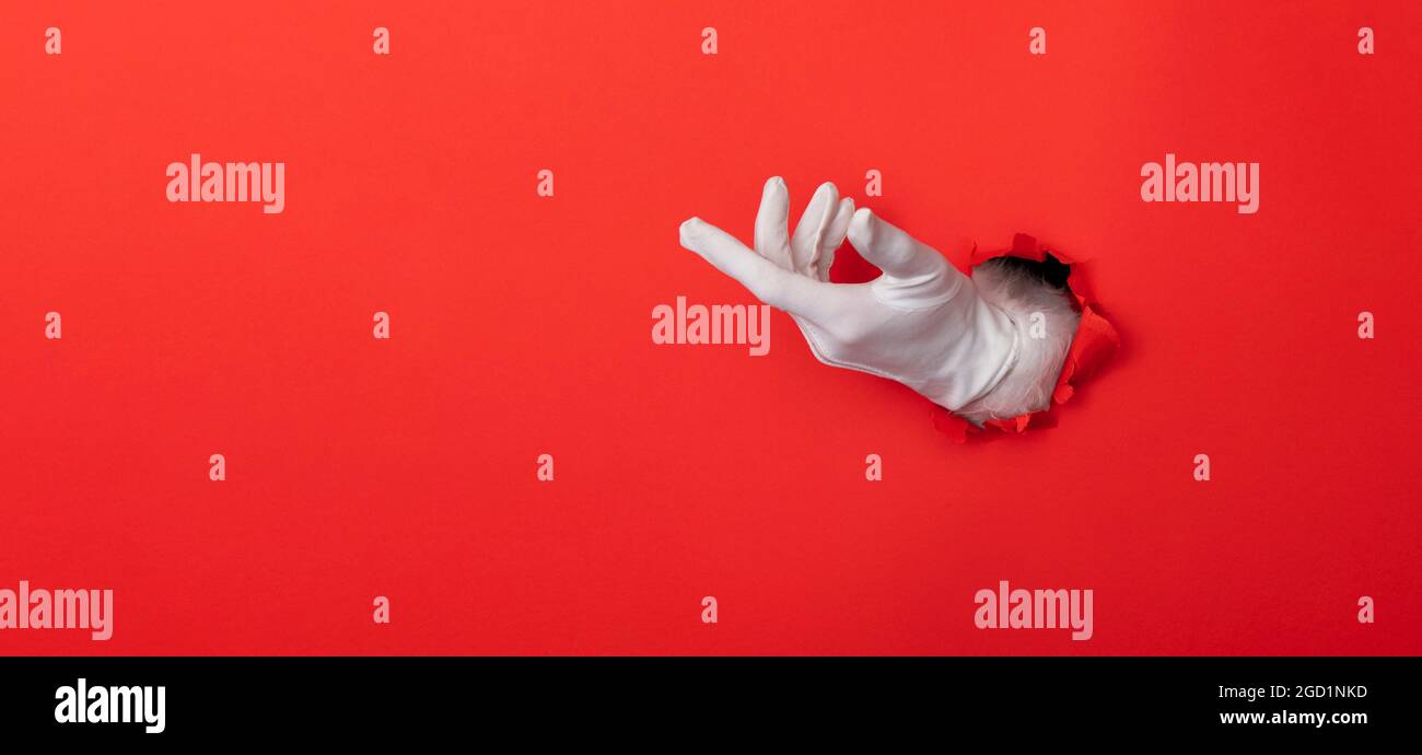 Main du Père Noël portant un gant blanc à travers un trou sur fond de papier rouge Banque D'Images