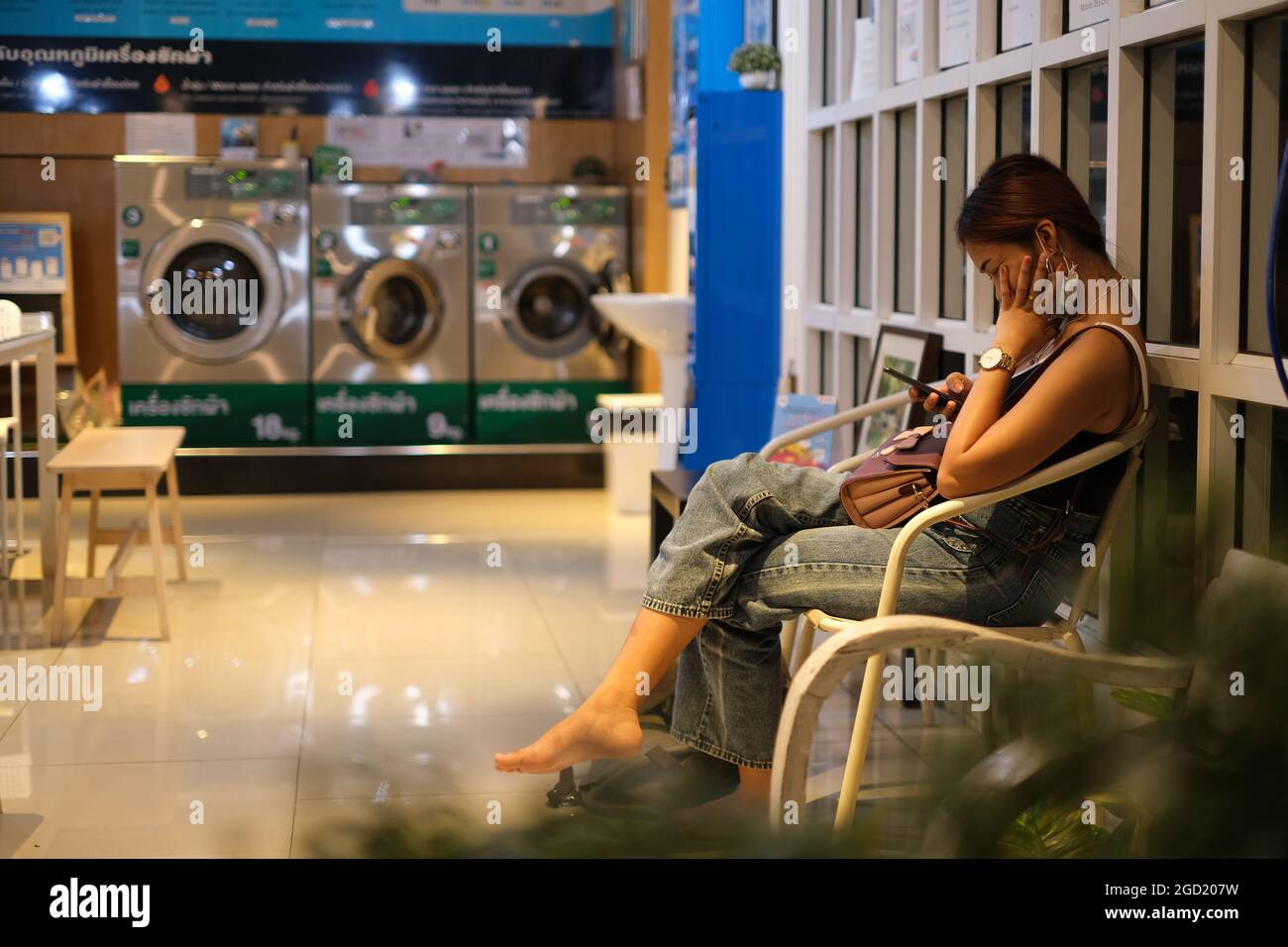 Une jeune femme thaïlandaise lit un smartphone en attendant dans un magasin de blanchisserie pour laver ses vêtements Banque D'Images