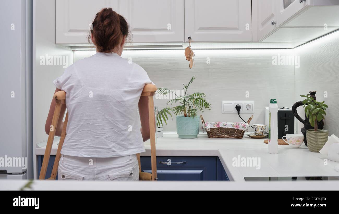 Femme sur des béquilles lave les plats dans la cuisine. Limitation temporaire des capacités physiques d'une personne dont la jambe est cassée. Phot de haute qualité Banque D'Images