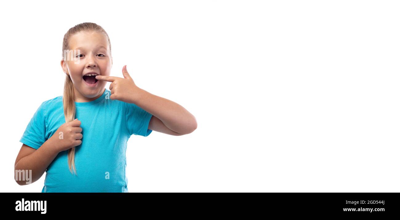 Une jeune fille d'âge préscolaire dans un T-shirt bleu montre un doigt à sa dent sur un fond blanc. La dent de lait est tombée, et une dent permanente pousse dans le TH Banque D'Images