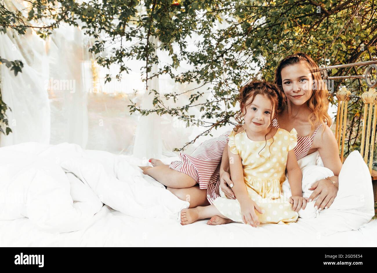 Jeune belle famille heureuse, mère souriante et petite fille assise sur le lit avec literie blanche propre dans le jardin vert à l'extérieur, maman et enfant Banque D'Images