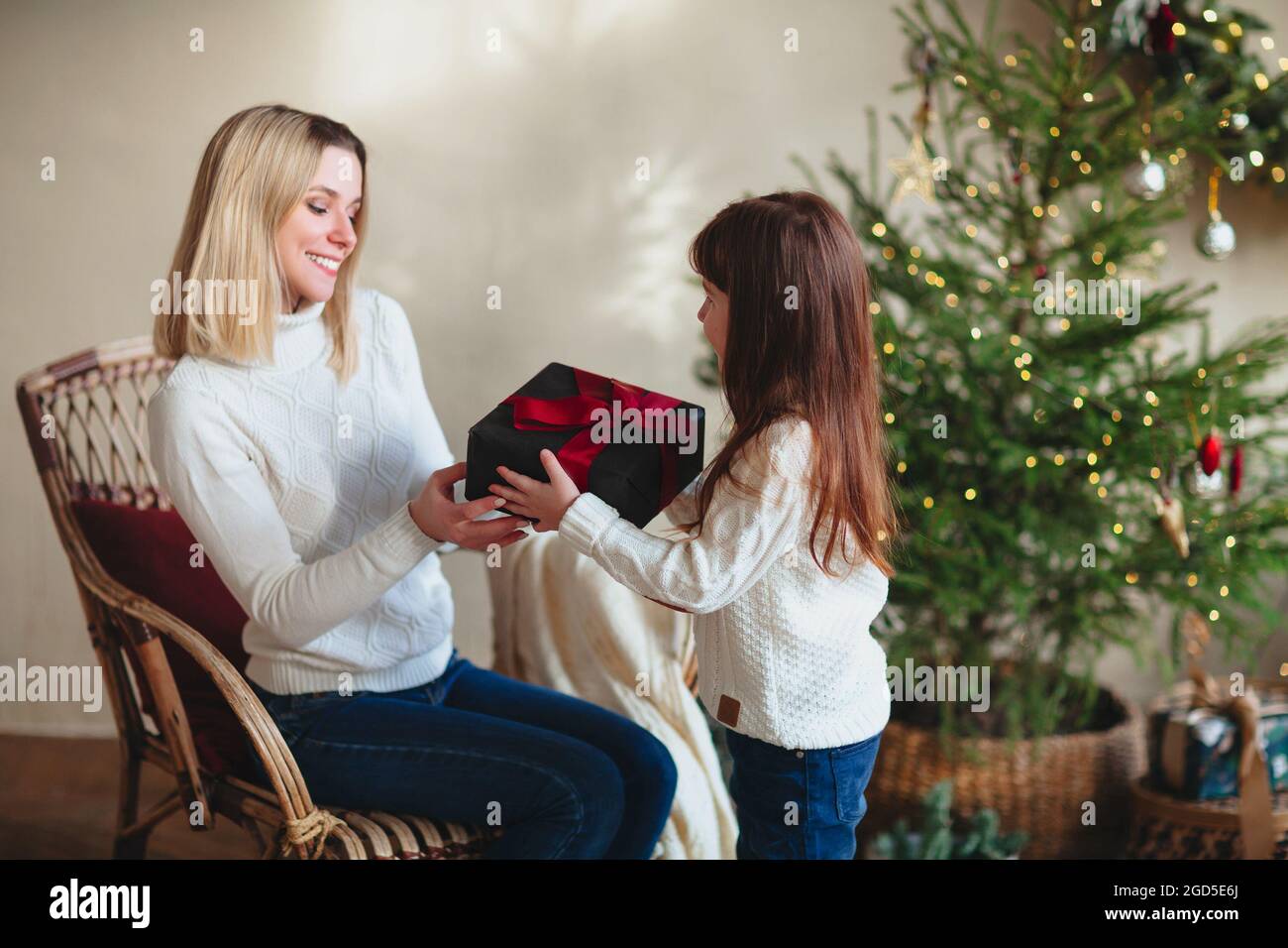 Fille mignonne cadeau de Noël sa jeune mère souriante, assise dans la chambre avec un arbre décoré, maman de famille heureuse et enfant portant un knitt blanc Banque D'Images