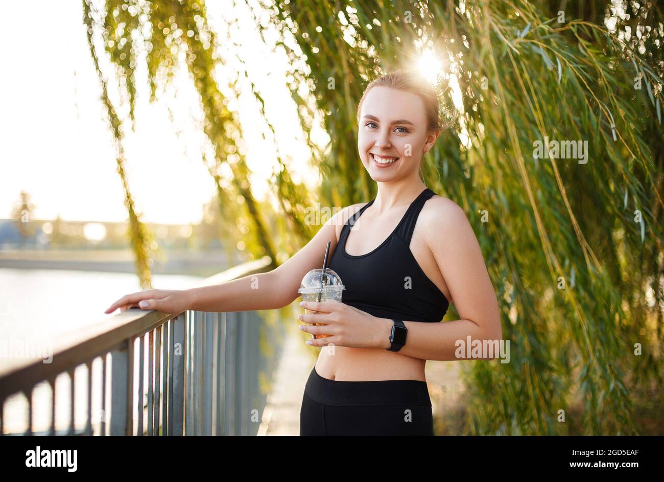 Jeune femme sportive en tenue sportive tenant la limonade à la main et souriant à la caméra tout en se tenant dans le parc près du lac, se reposant après une course matinale à la nat Banque D'Images