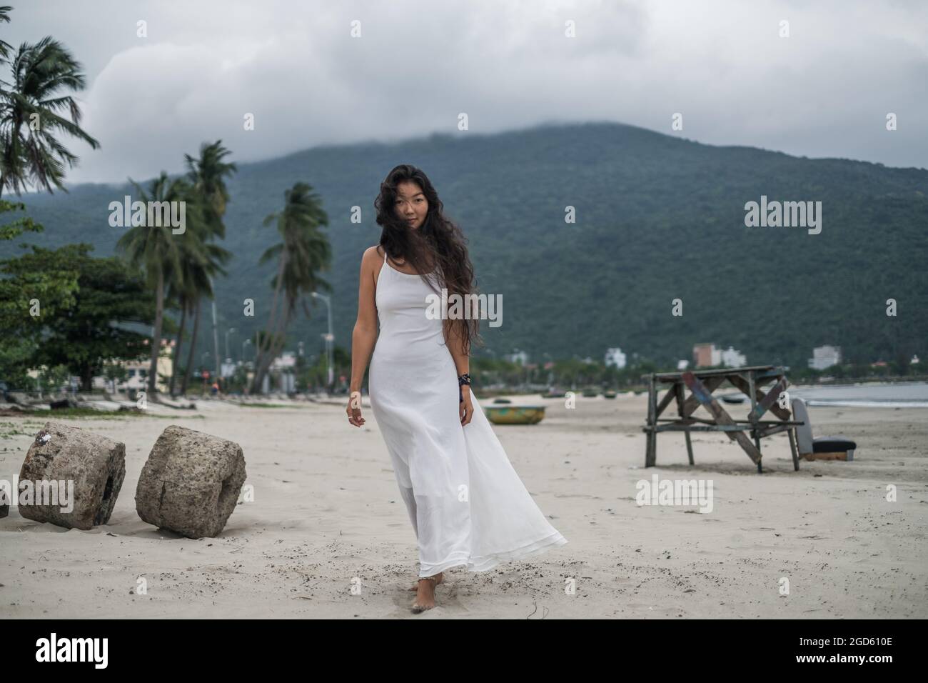 Belle jeune femme asiatique en robe blanche marchant sur la plage. Fond de montagne. Cheveux longs et bouclés noirs. Vent dans ses cheveux. Photo romantique. Banque D'Images
