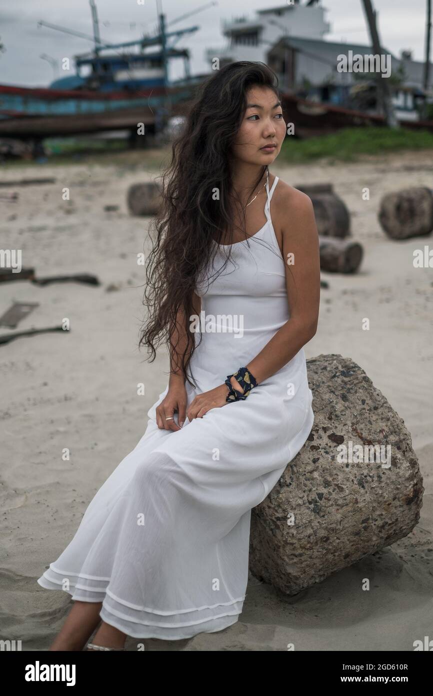 Charmante jeune femme asiatique en robe blanche assise sur la pierre et regardant de côté. Brunette cheveux longs bouclés. Photo romantique. Photo de haute qualité Banque D'Images