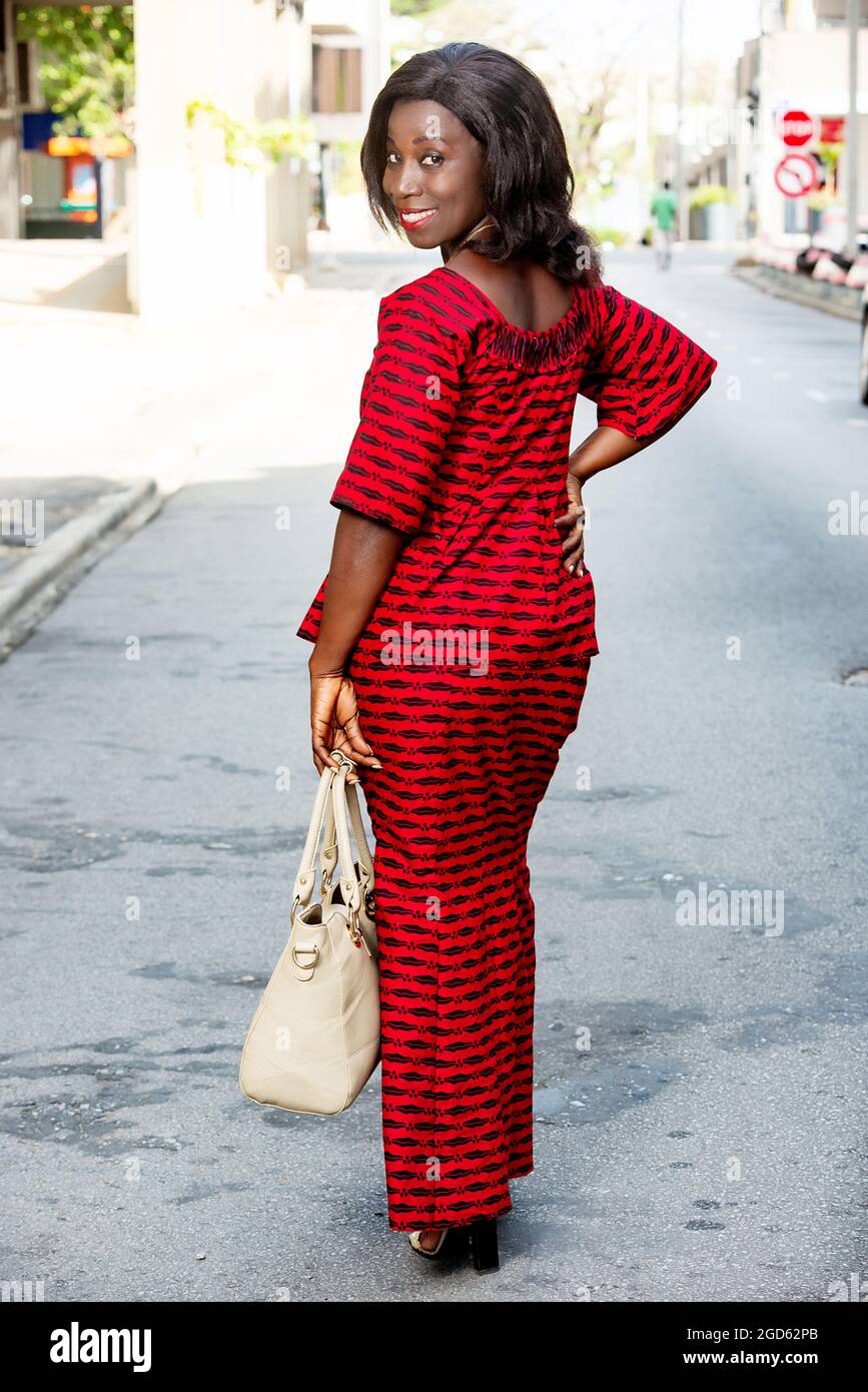 Femme africaine debout dans la rue et regardant en arrière la caméra souriante. Femme concept en tissu rouge ou de mode africaine Banque D'Images