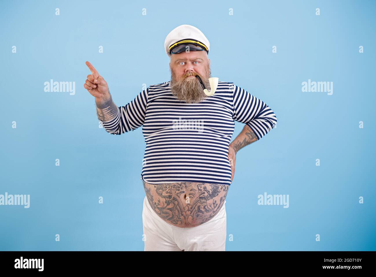 Homme marin de plus grande taille avec tatouages et fumer des points de tuyau de côté par le doigt sur fond bleu clair Banque D'Images