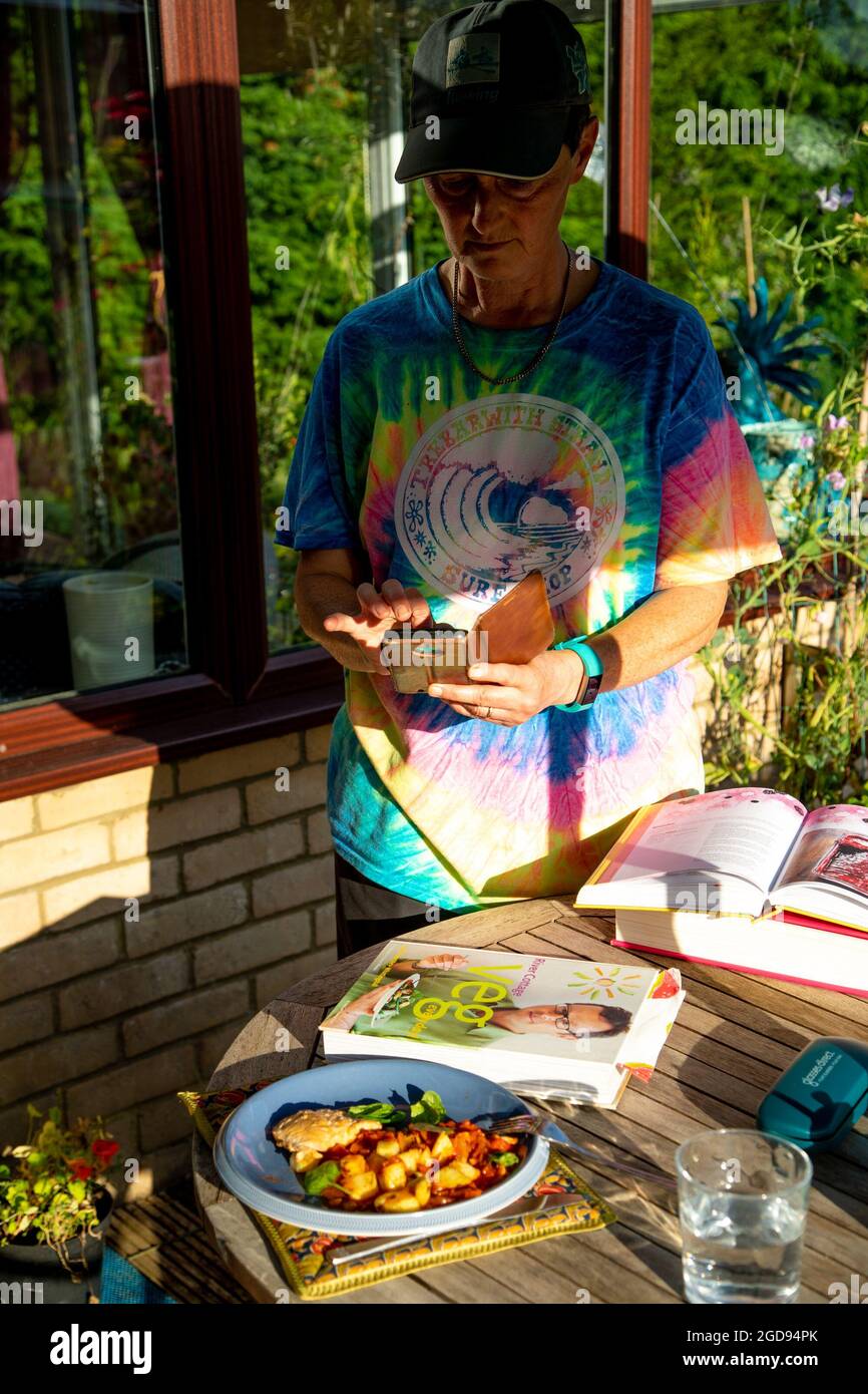 Une femme d'âge moyen prenant une photo d'une assiette de nourriture colorée. Il y a des livres de cuisine sur la table. Banque D'Images
