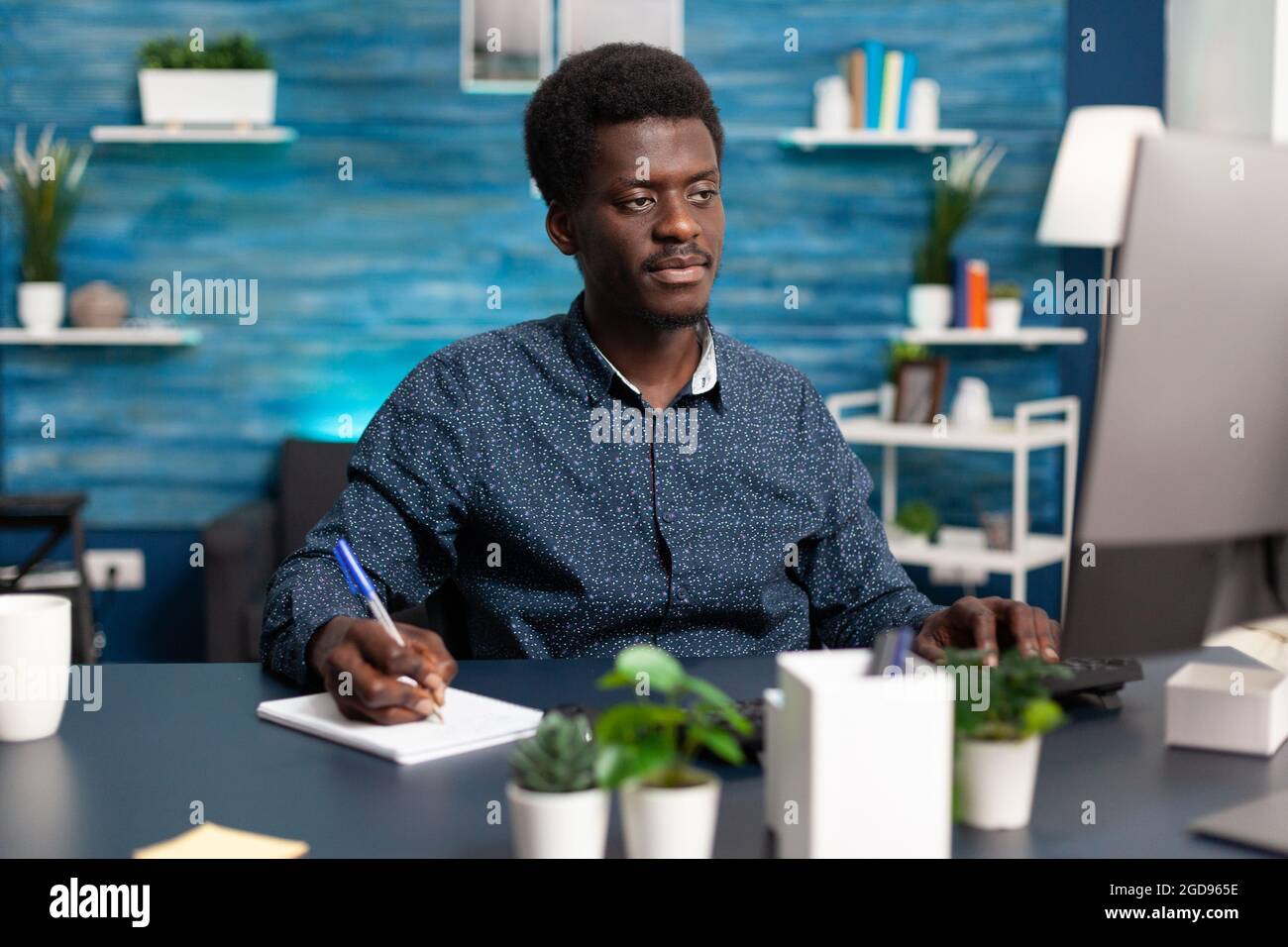 Indépendant prenant des notes sur ordinateur portable regardant l'ordinateur sur le bureau à domicile. Afro-américain homme travaillant à distance à la maison et faisant des cours d'université en ligne pour les affaires de médias sociaux Banque D'Images