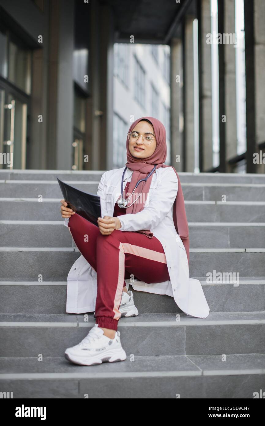 Jolie femme musulmane asiatique médecin, portant hijab, des gommages rouges  et un manteau blanc, assis sur des escaliers modernes gris à l'extérieur du  centre médical et d'urgence, tenant la tomographie par rayons