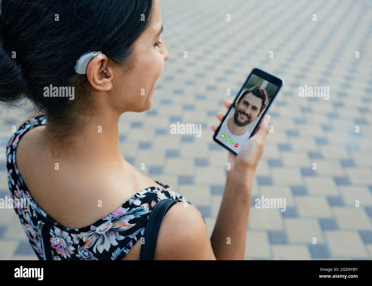 La femme Brunette, avec une prothèse auditive derrière l'oreille, communique avec son petit ami par communication vidéo via un smartphone Banque D'Images