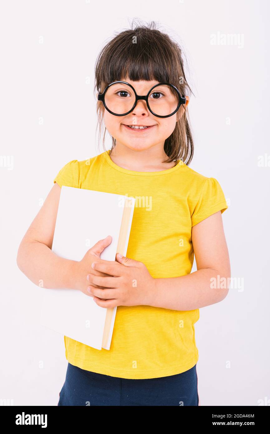 Petite fille souriante portant un t-shirt jaune et des lunettes noires rondes tenant un livre dans ses mains sur fond blanc Banque D'Images