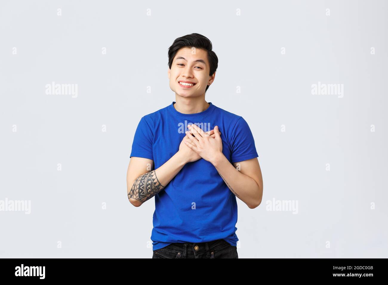 Touché et reconnaissant beau mâle asiatique en bleu t-shirt avec tatouages, touchant le coeur et sourire, apprécier l'aide, être heureux ou ravie Banque D'Images