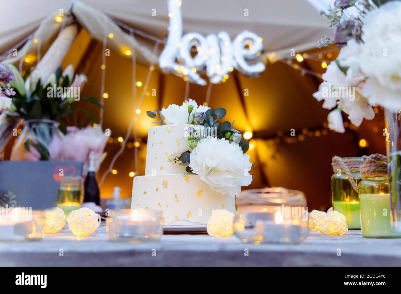 Gâteau de mariage à deux niveaux décoré de fleurs de pivoine fraîches et d'or comestible sur la table en bois avec fleurs, bougies. Mariage en extérieur avec lumières Banque D'Images