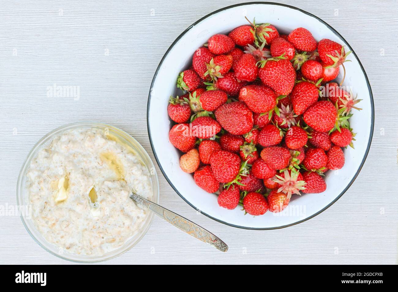 De délicieuses fraises mûres fraîches dans un bol en métal blanc et une assiette de porridge de flocons d'avoine avec beurre sur une table blanche. Banque D'Images