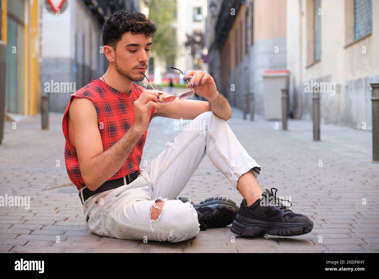 Jeune homme caucasien avec de longs ongles faux tenant des lunettes de soleil assis sur le sol. Banque D'Images