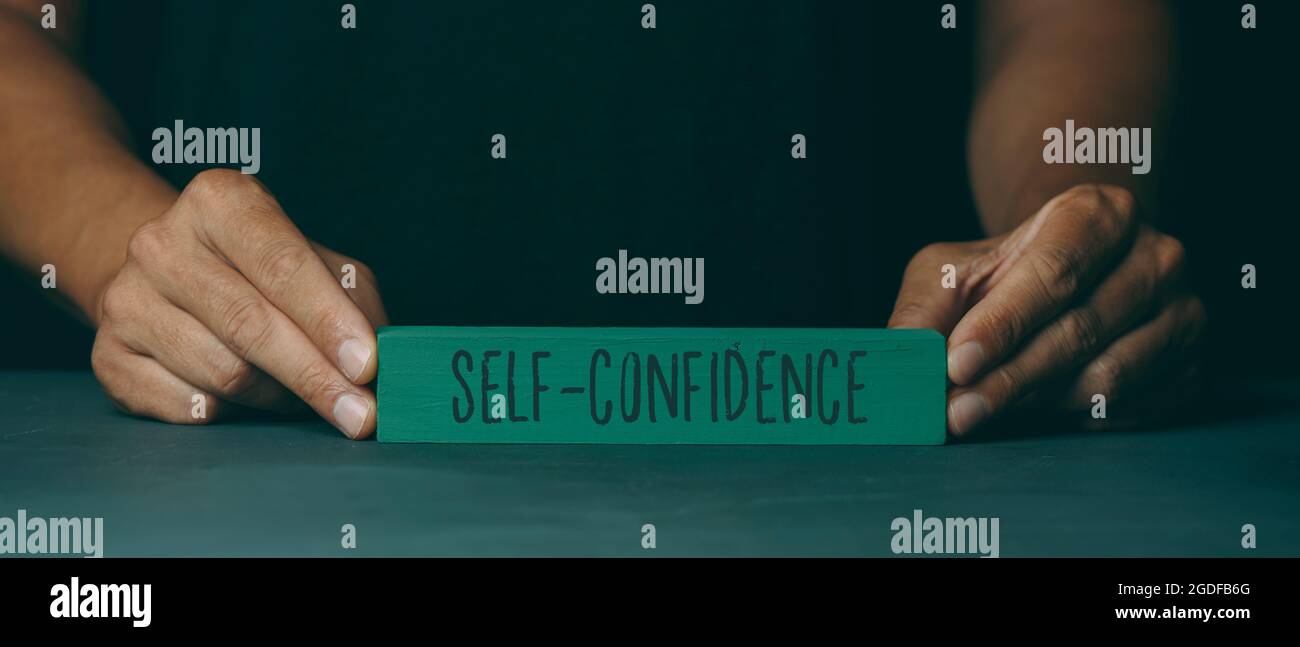 un homme caucasien montre un bloc de construction vert avec le texte de confiance en soi écrit, sur une surface gris foncé, dans un format panoramique à utiliser comme web Banque D'Images