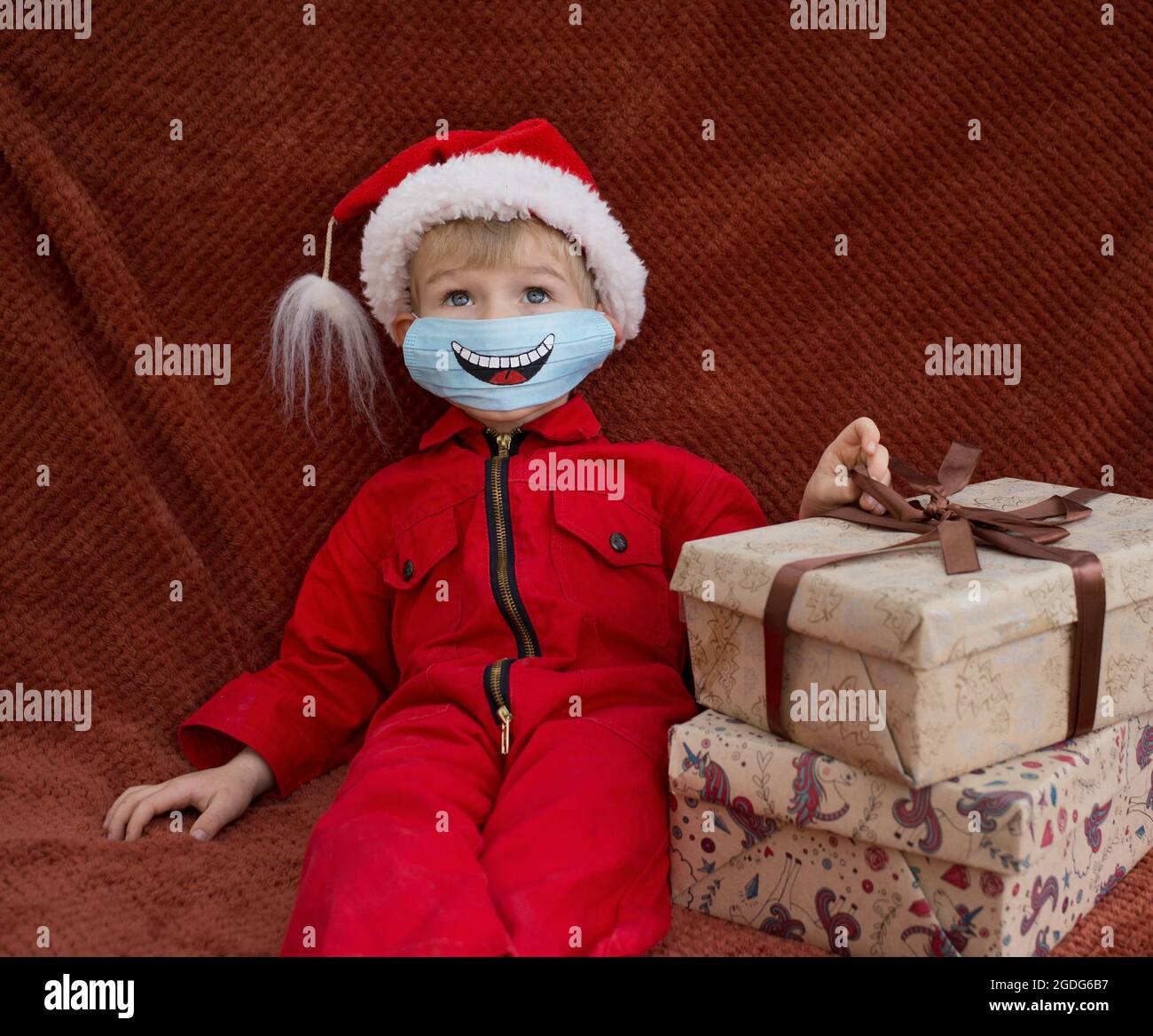 Enfant Dans Une Combinaison Rouge Le Chapeau De Pere Noel Se Trouve Pres Des Boites Cadeaux Sur Le Visage Il Y A Un Masque Medical Bleu Avec Le Sourire Rigolo Peint Il Est
