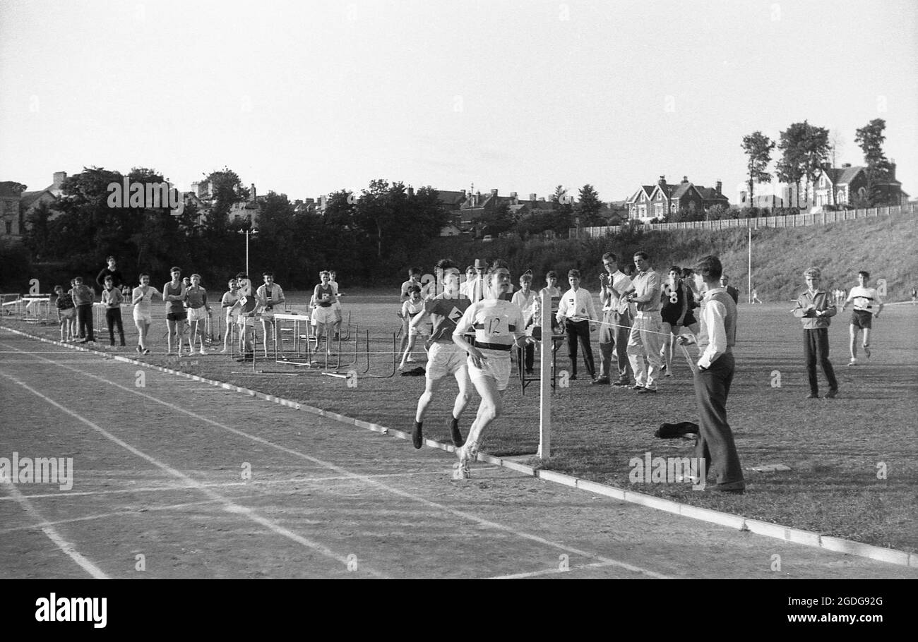 1964, historlcal, à l'extérieur d'une journée de sport dans les écoles de comté, une fin de près sur la piste de Cinder comme deux écoliers course pour la bande d'arrivée, tenue par un jeune homme sur le côté de la piste, Exeter, Devon, Angleterre, Royaume-Uni. Banque D'Images