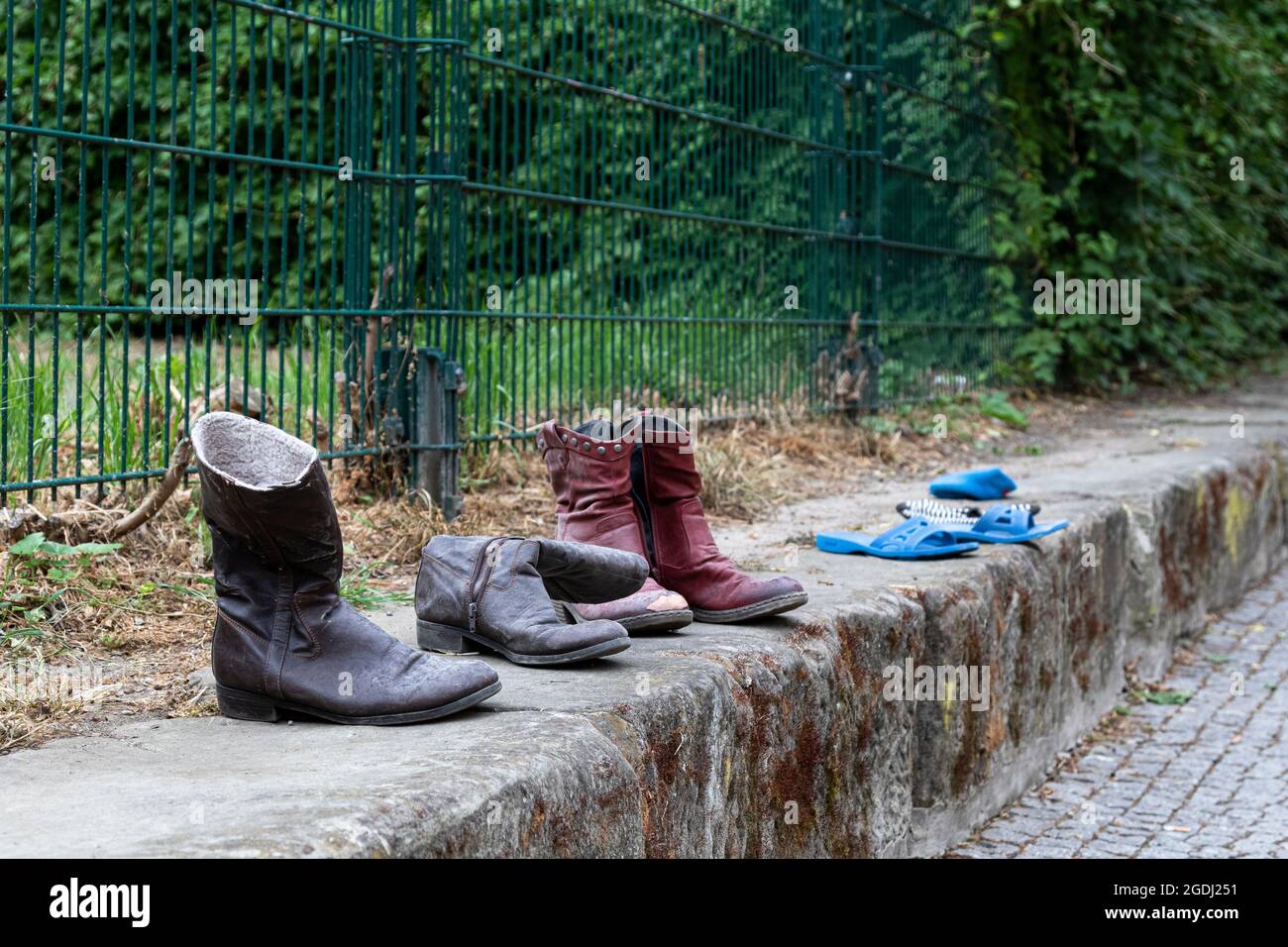 Vieilles bottes et sandales alignées sur un mur bas avec une clôture en métal vert en arrière-plan. Banque D'Images