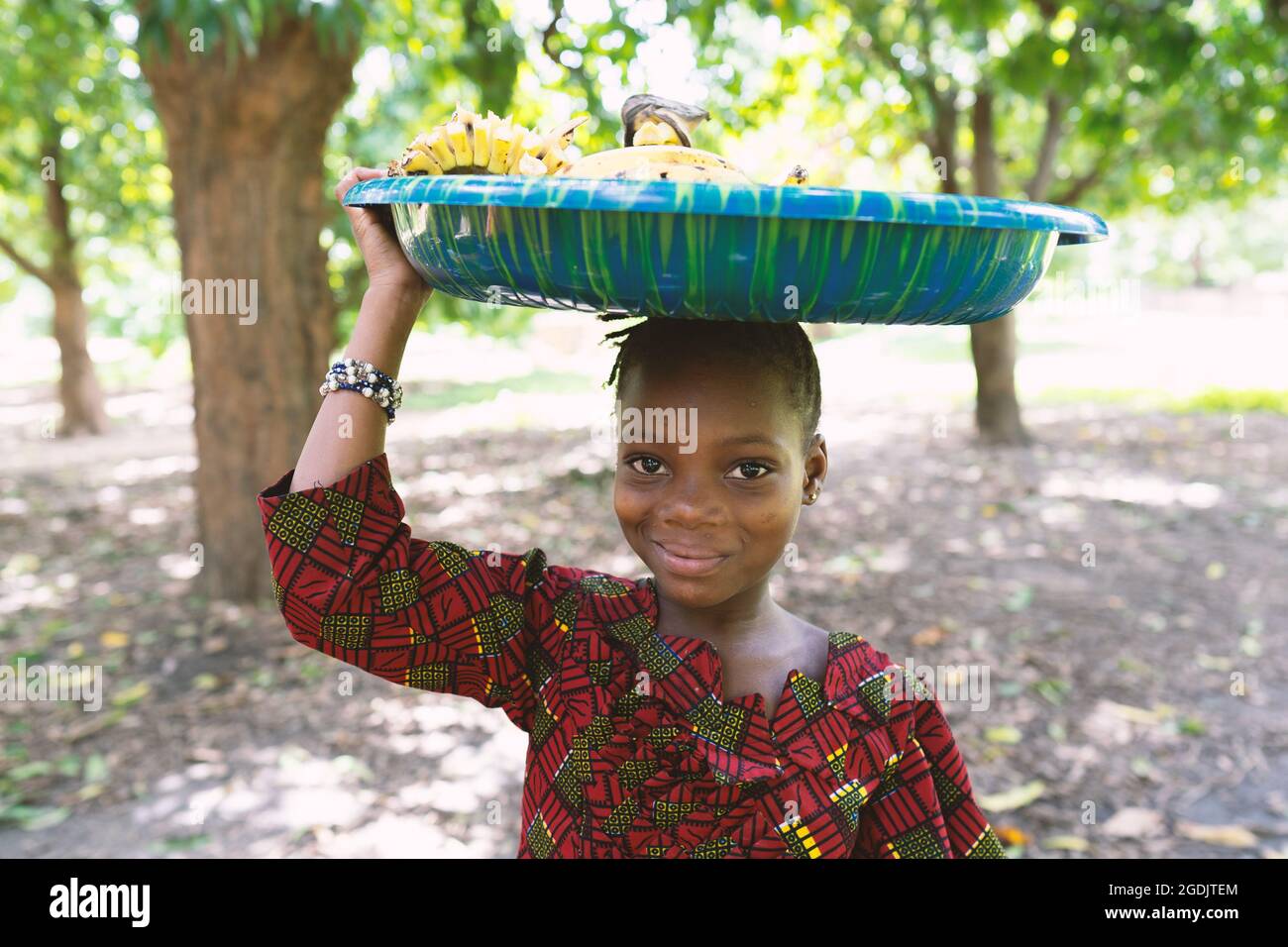 Sourire belle fille noire portant une grande plaque en plastique bleu sur sa tête pleine de bananes Banque D'Images