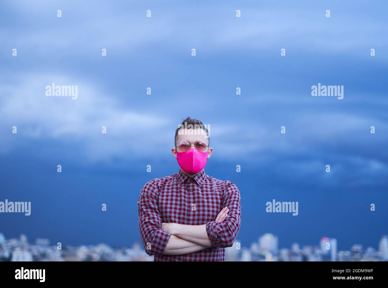 Mignon caucasien gay dans le masque de protection rose et les lunettes de soleil roses regardant directement dans l'appareil photo. Concept d'espoir, thème LGBTQ. Portrait de homme avec fond de temps pluvieux et horizon urbain Banque D'Images