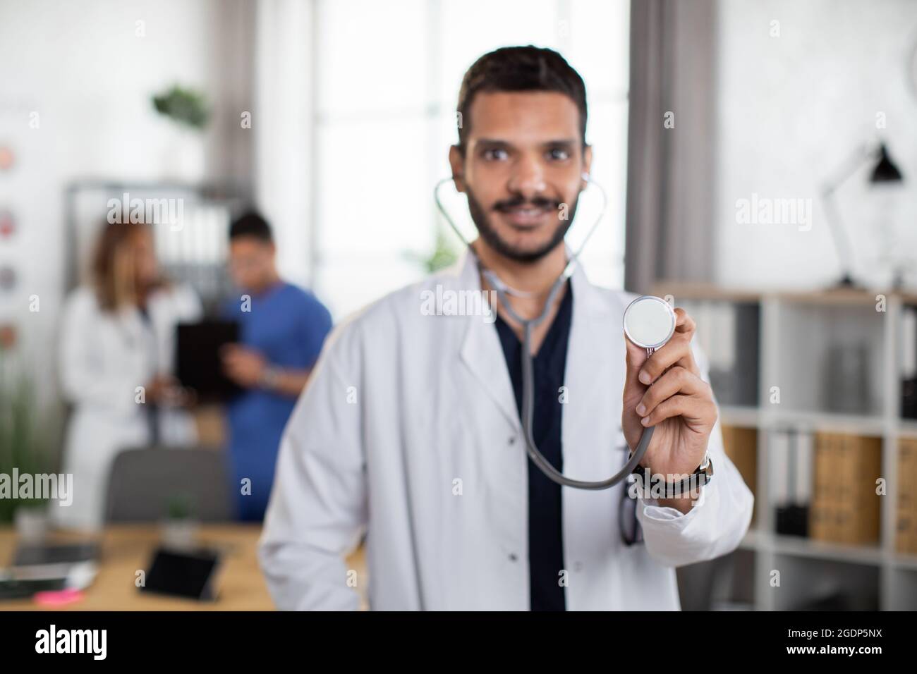 Vue rapprochée floue d'un beau médecin indien, debout avec un stéthoscope dans une salle d'embarquement moderne et légère, avec deux collègues différents parlant en arrière-plan. Concentrez-vous sur le stéthoscope Banque D'Images