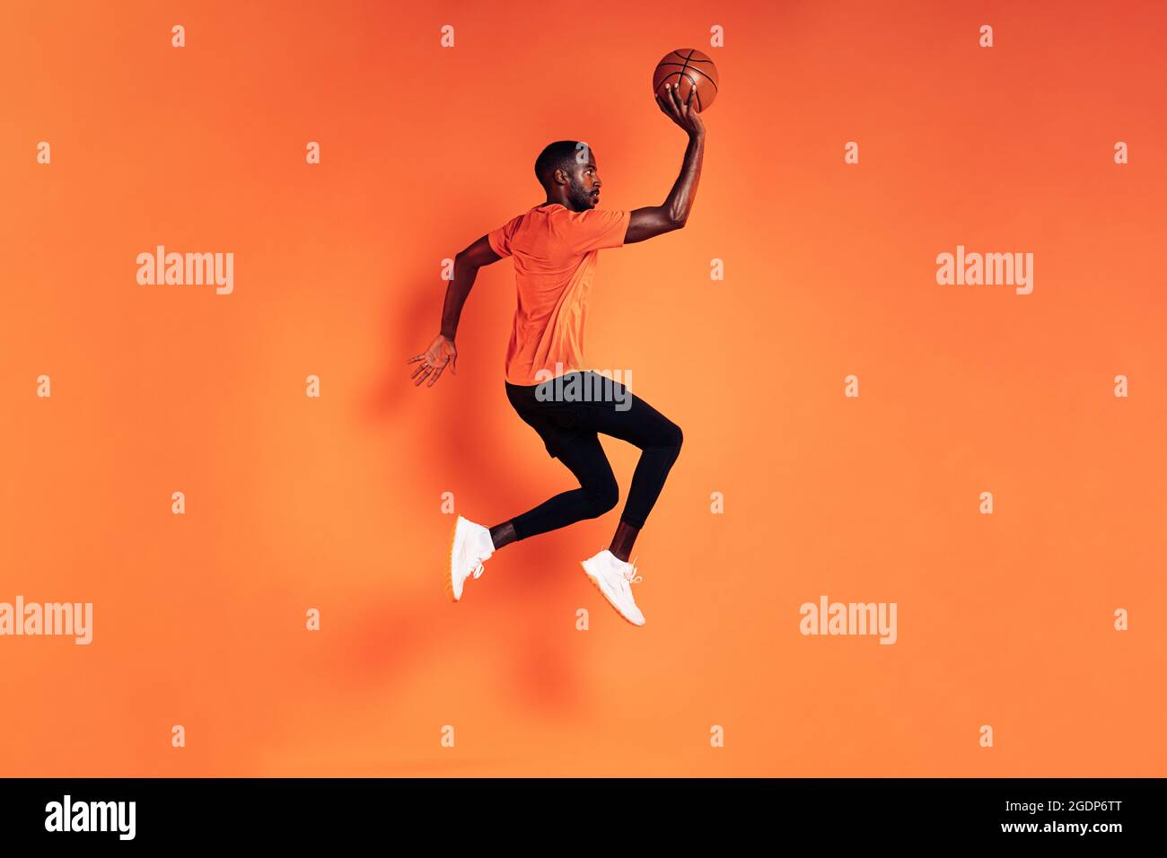 Athlète masculin sautant avec le ballon de basket. Vue latérale du sportif s'exerçant sur fond orange. Banque D'Images