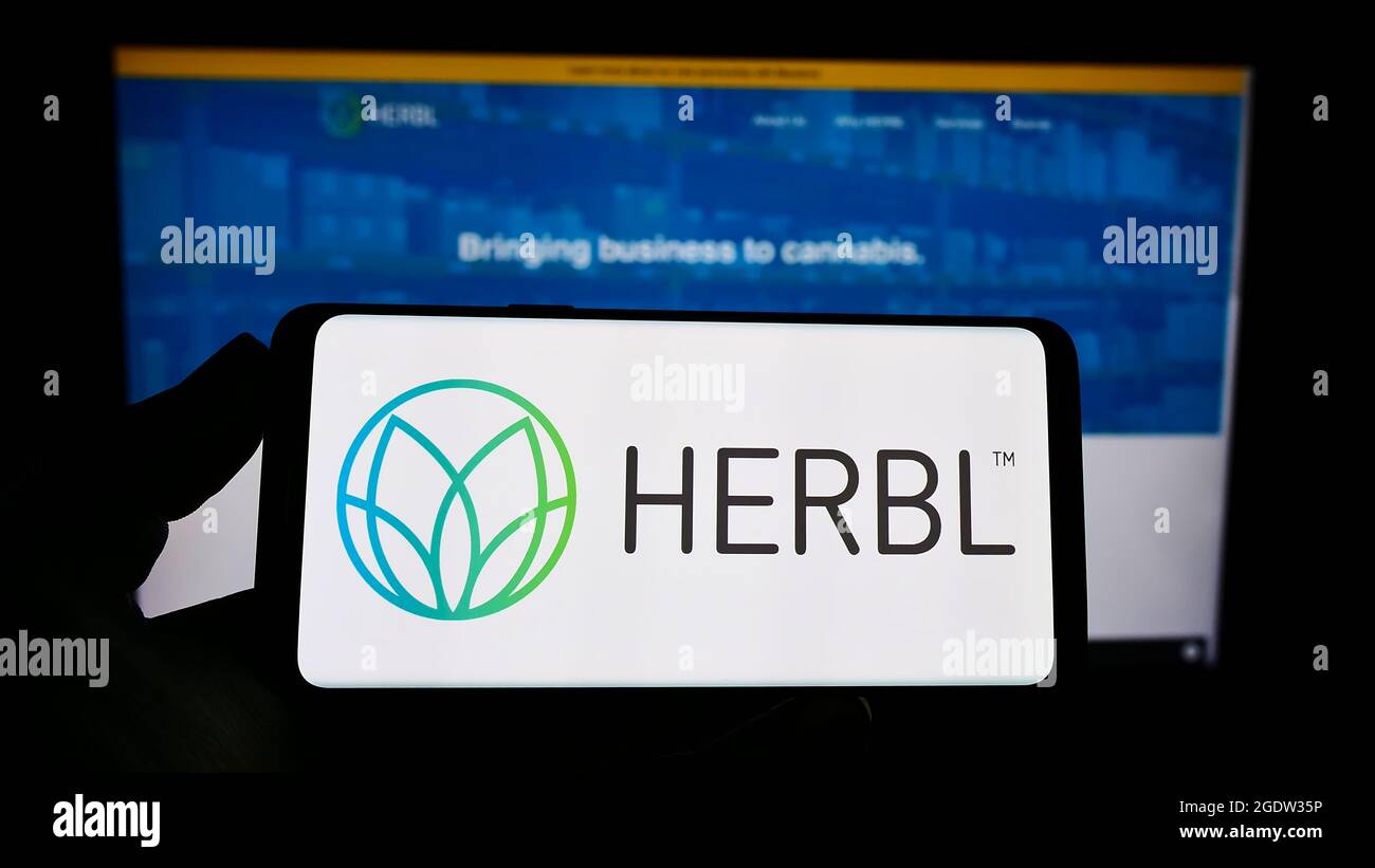 Personne tenant un téléphone portable avec le logo de la société américaine HERBL de la chaîne d'approvisionnement en cannabis sur l'écran en face de la page Web d'affaires. Mise au point sur l'affichage du téléphone. Banque D'Images