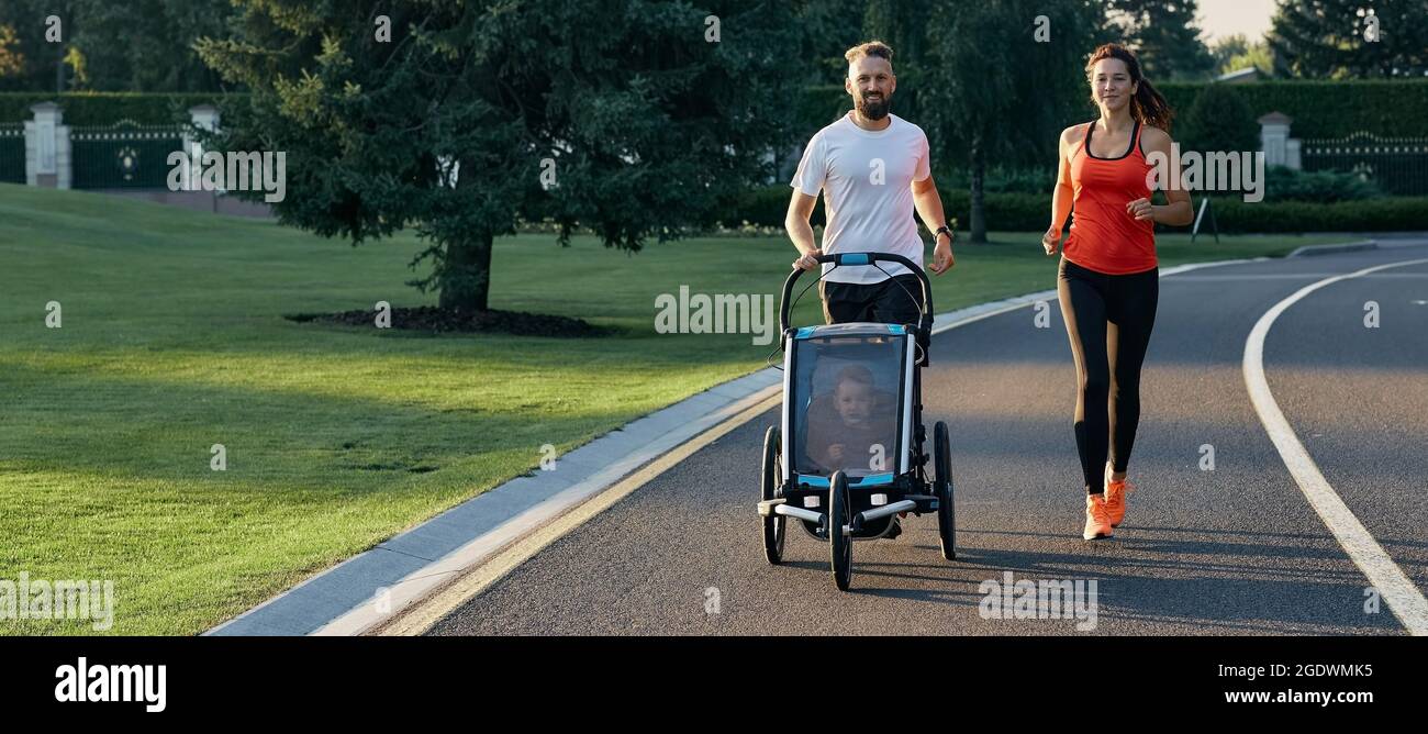 Une jeune famille avec son enfant dans une poussette de jogging pendant le jogging dans un parc public. Famille active courant Banque D'Images