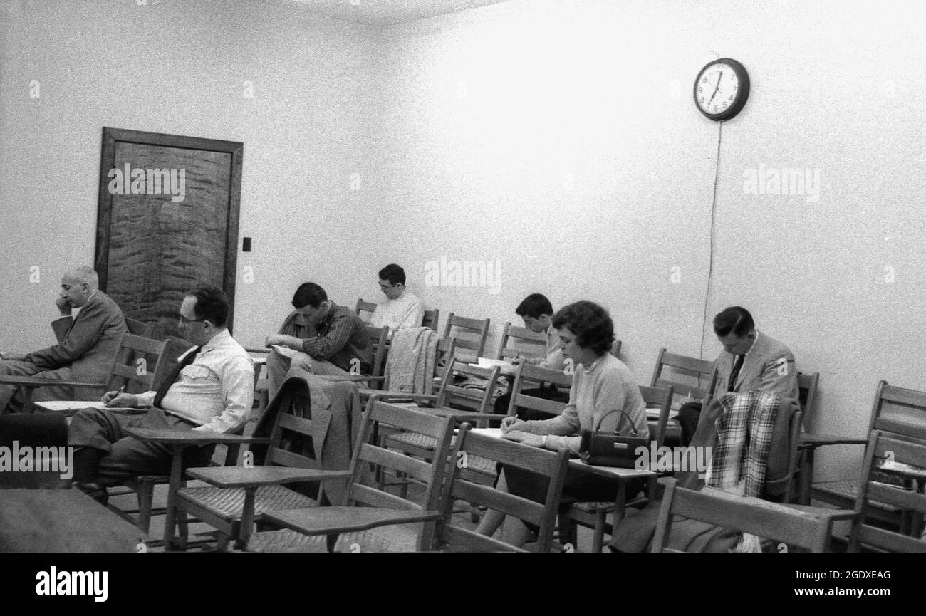 Années 1960, histoire, études complémentaires, lors d'un cours du soir, un petit groupe d'étudiants matures assis dans une classe faisant des notes écrites sur les chaises-bureaux traditionnels en bois de l'époque, Etats-Unis. Les bureaux d'école ont une histoire intéressante et la vieille école, ensemble chaise-bureau non réglable vu ici, était juste l'un des nombreux styles de bureau qui ont été introduits dans la salle de classe au fil des ans. Banque D'Images