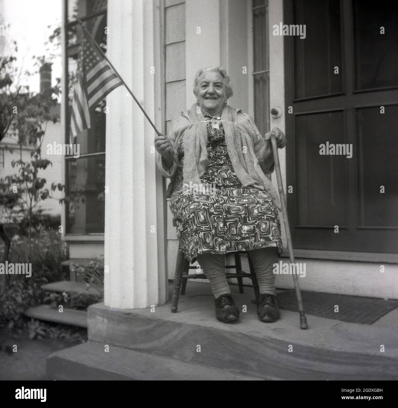 Années 1950, historique, une fière américaine... une dame âgée assise sur une chaise en bois sur un pas en pierre devant sa porte d'entrée, avec une baguette dans une main et dans l'autre, le drapeau américain, États-Unis. Avec ses 50 étoiles et ses 13 bandes, le drapeau est le symbole puissant de la nation américaine, Banque D'Images