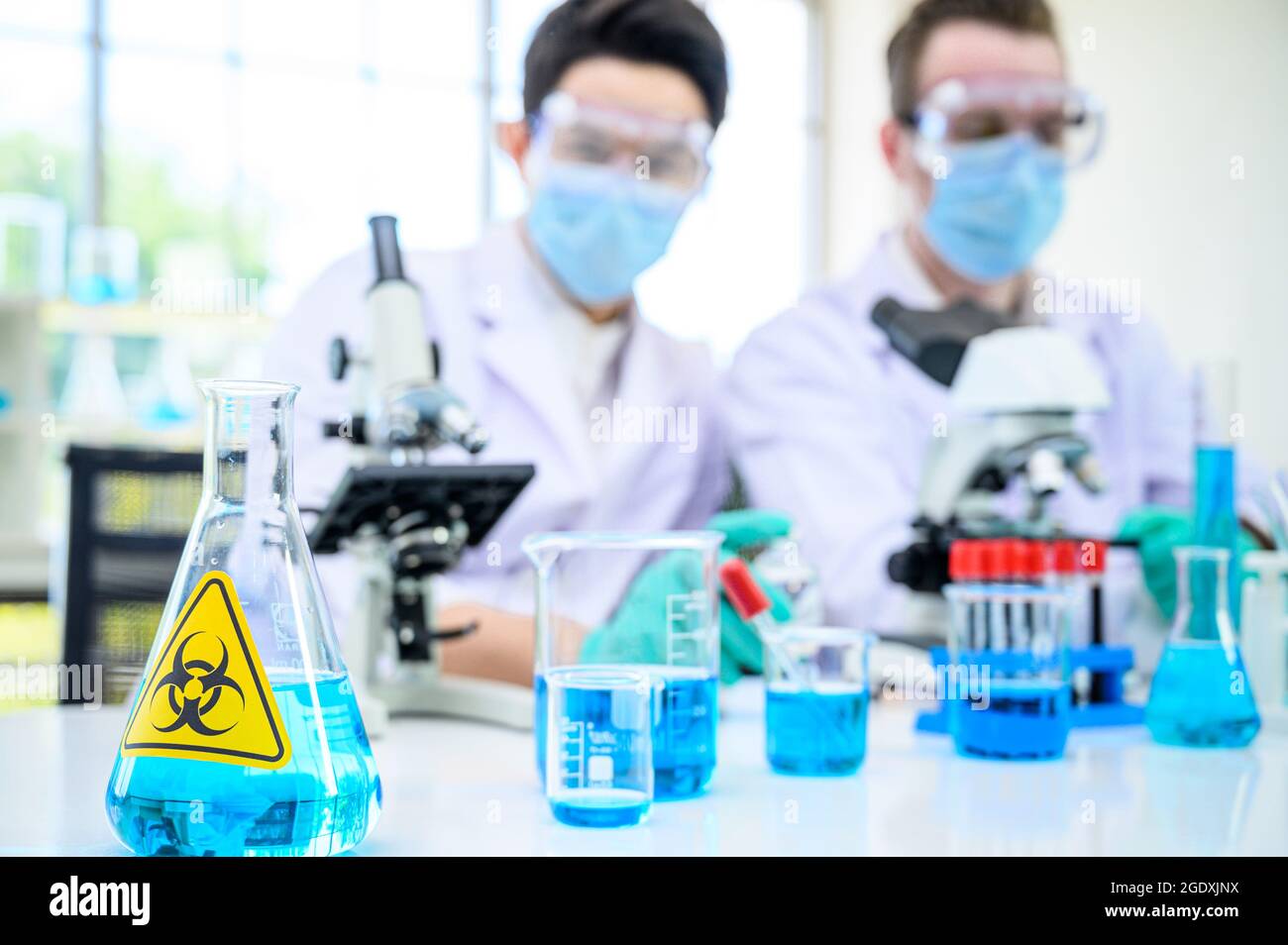 Équipement de laboratoire avec liquide bleu à l'intérieur du support sur la table avec couple scientifique mâle portant une combinaison de protection travaillant avec un microscope pour la recherche à l Banque D'Images