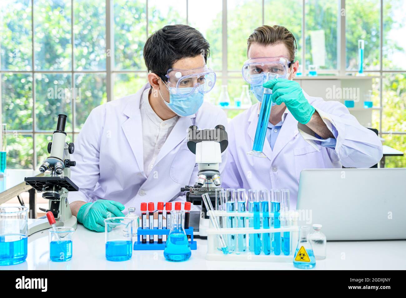 Couple scientifique mâle portant une combinaison de protection tenant le bécher ou le tube avec un échantillon de liquide à l'intérieur et en travaillant avec de nombreux équipements de laboratoire pour la recherche en laboratoire Banque D'Images