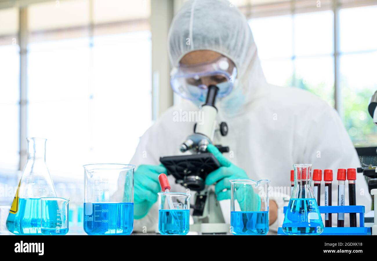 Équipement de laboratoire avec liquide bleu à l'intérieur du statif sur la table avec un scientifique mâle portant une combinaison de protection travaillant avec le microscope pour la recherche du coronavirus Banque D'Images