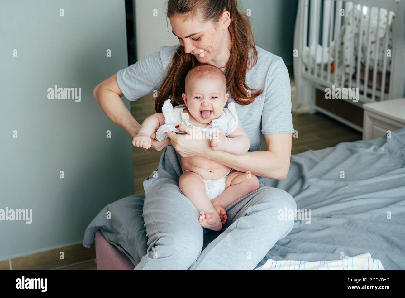 Une jeune mère habille son bébé gai assis sur ses genoux et montrant la langue. Banque D'Images