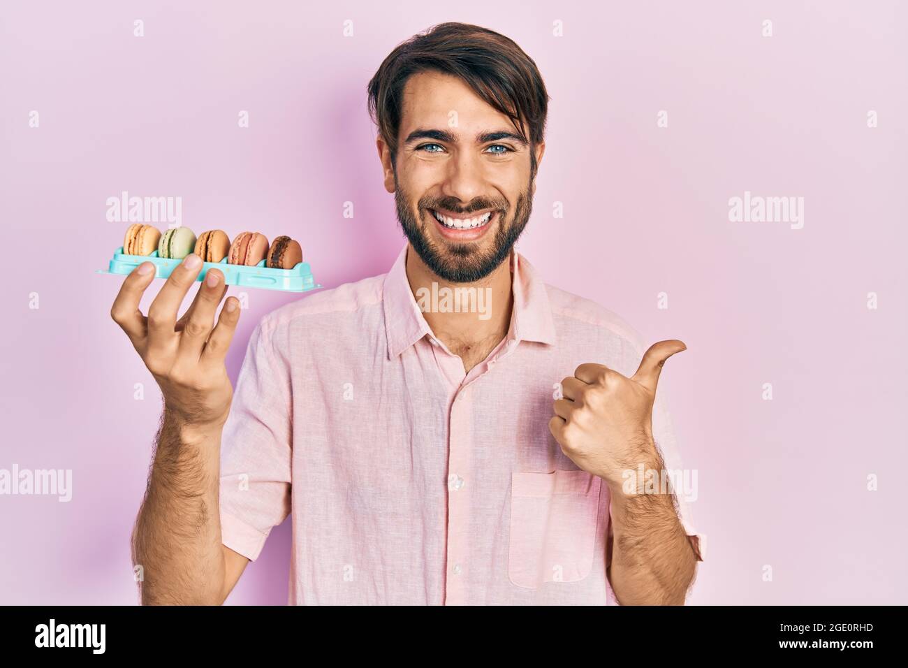 Jeune homme hispanique portant de délicieuses pâtisseries de macrons pointant le pouce vers le côté souriant heureux avec la bouche ouverte Banque D'Images
