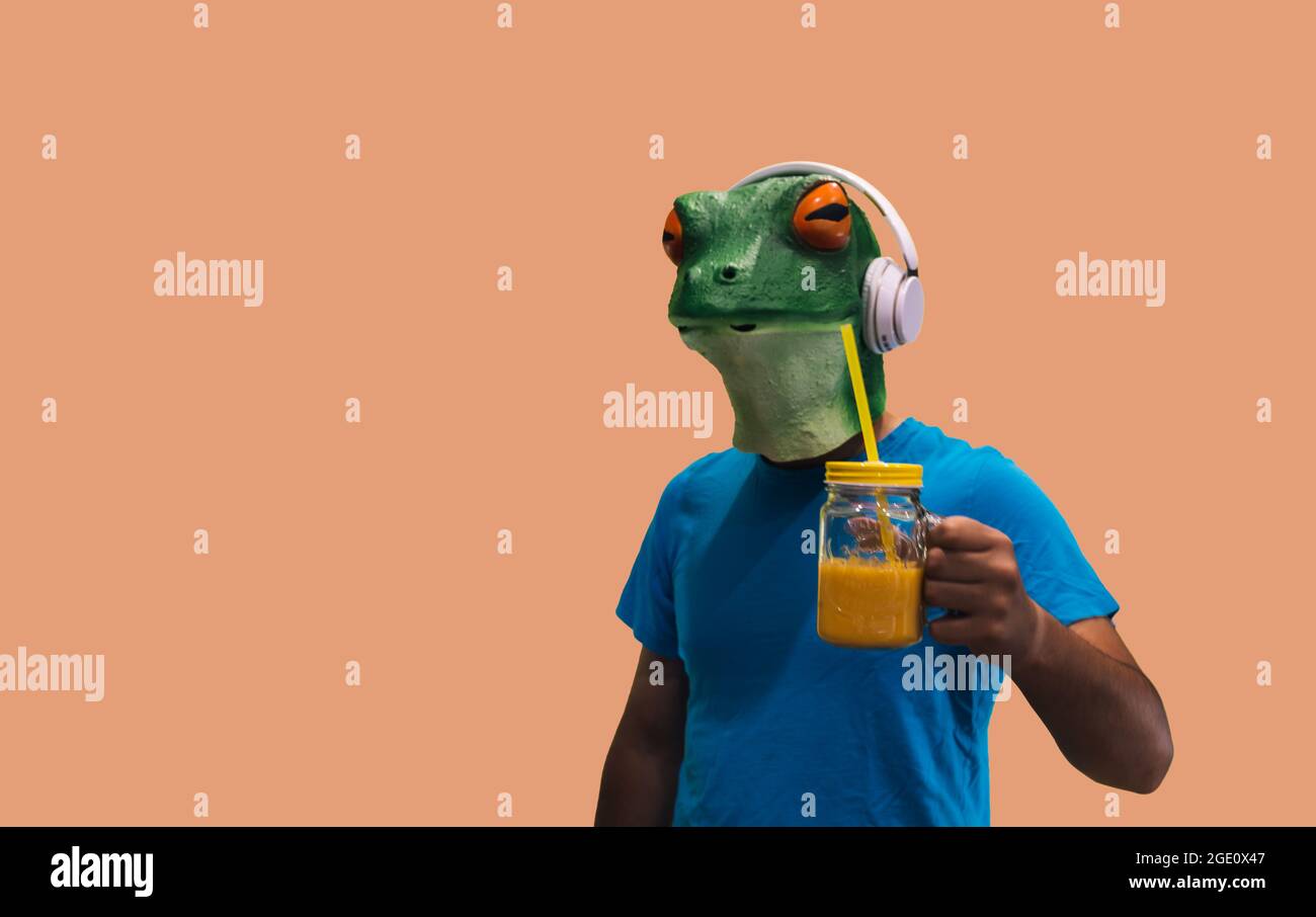 personne avec un masque de grenouille avec un pot en verre avec du jus de fruits et une paille à boire et à rafraîchir tout en écoutant de la musique à travers des écouteurs sur le backgro orange Banque D'Images