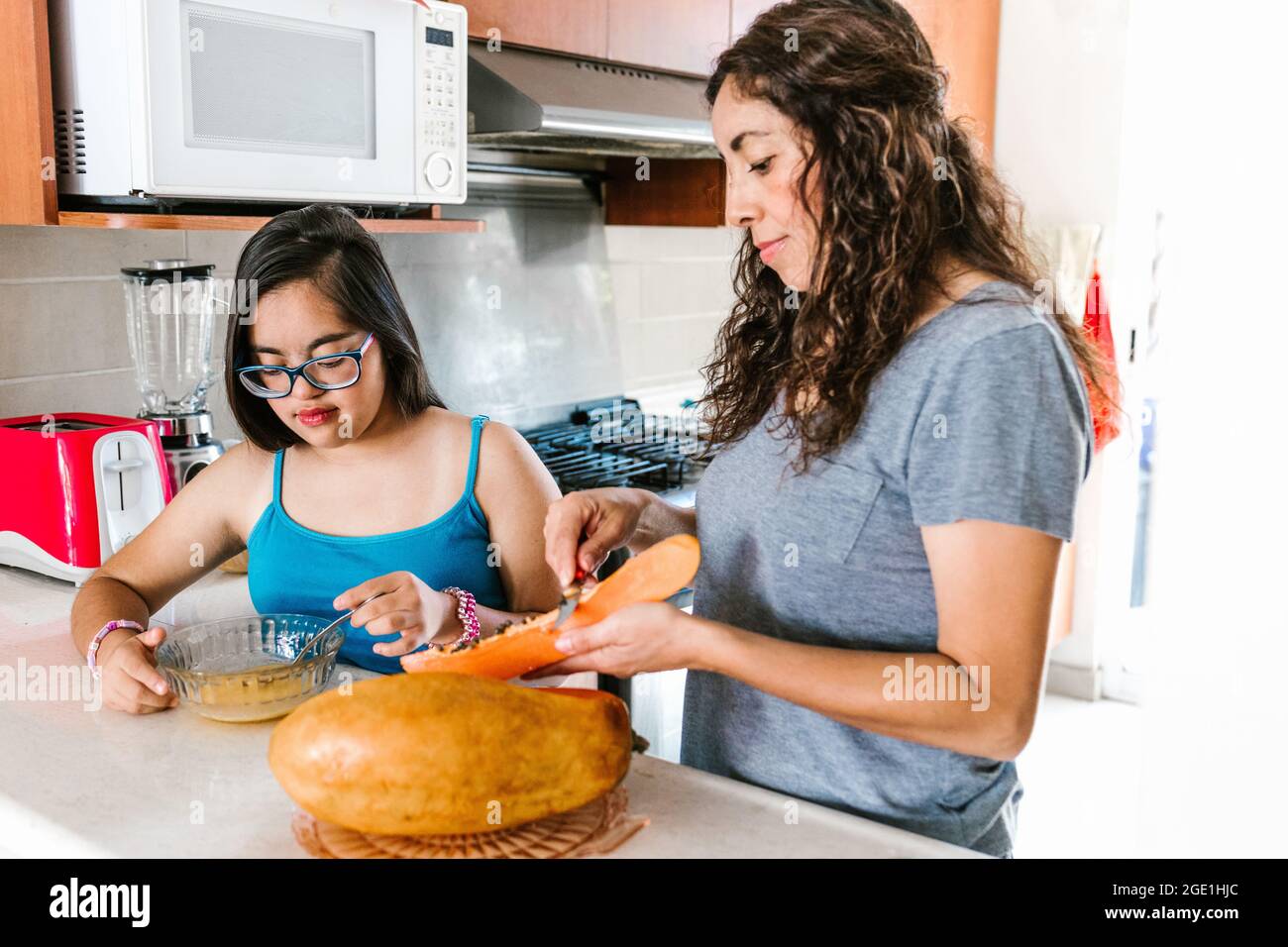 Une maman latine trancheuse des fruits avec sa fille adolescente avec le syndrome de Down dans la cuisine, dans le concept de handicap en Amérique latine Banque D'Images