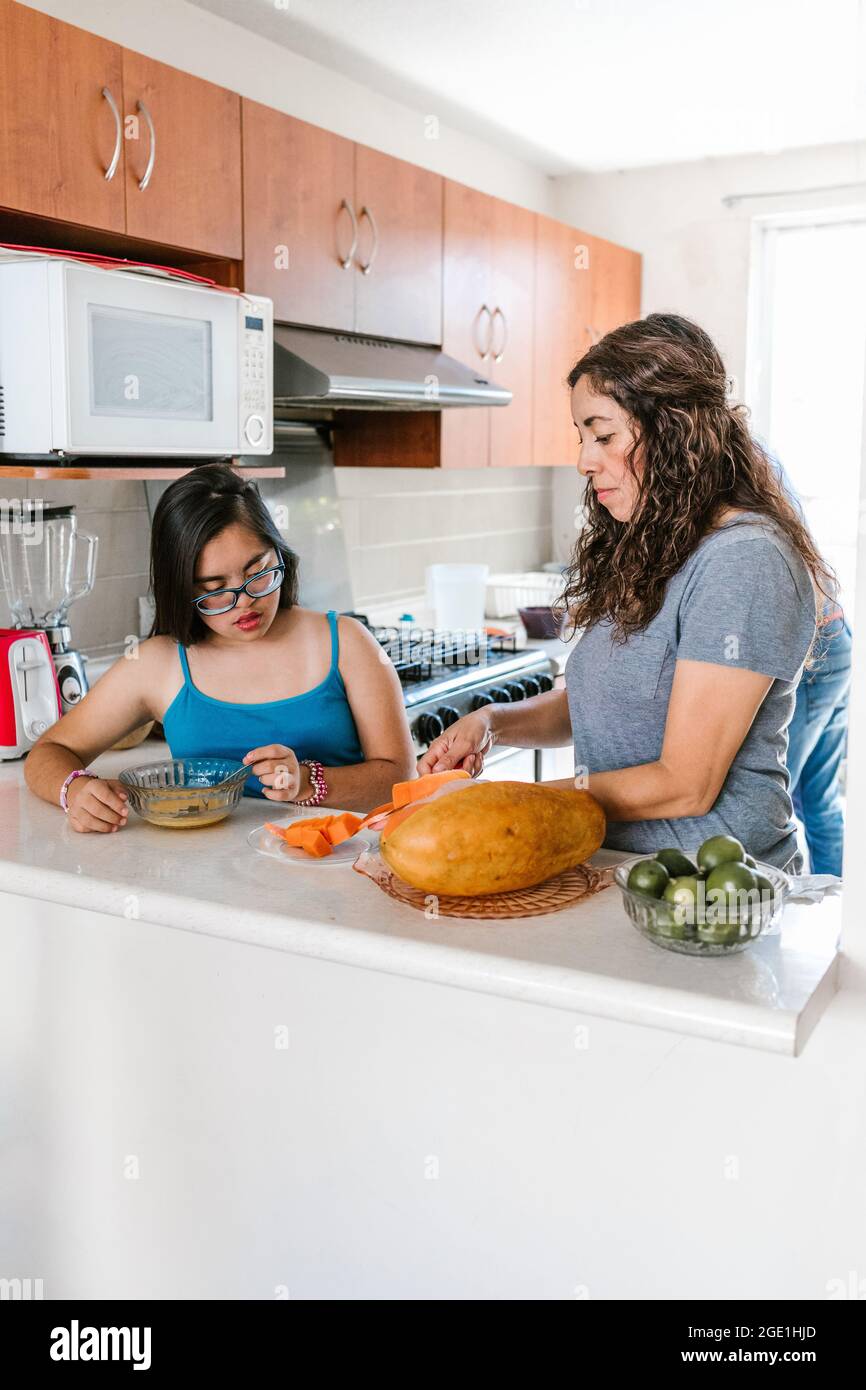 Une maman latine trancheuse des fruits avec sa fille adolescente avec le syndrome de Down dans la cuisine, dans le concept de handicap en Amérique latine Banque D'Images