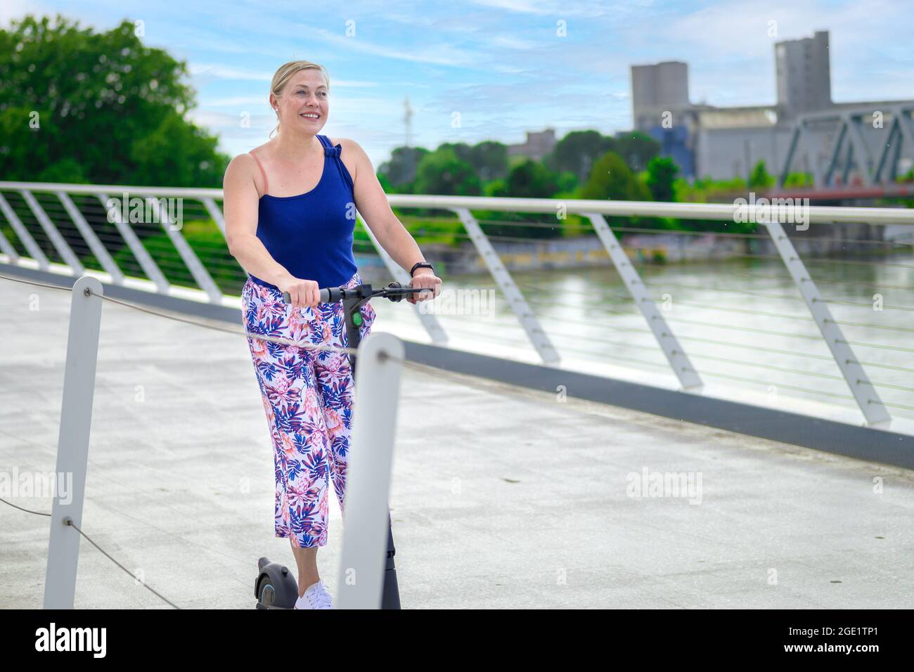 Femme de souche d'âge moyen à bord d'un scooter électrique traversant un pont au-dessus d'une rivière dans une ville dans un concept de transport urbain et de mobilité Banque D'Images